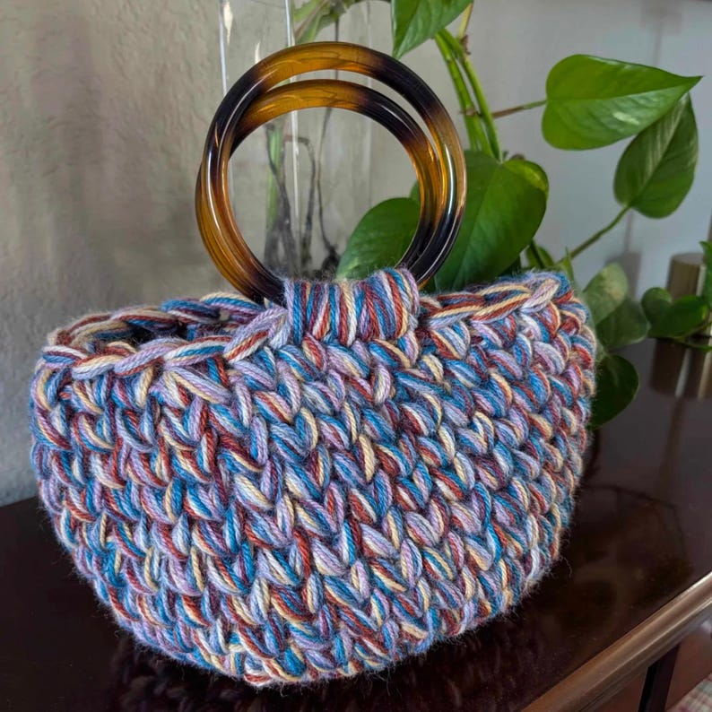 May include: A handmade crochet bag with a round shape and a tortoiseshell handle. The bag is made of colorful yarn in shades of blue, purple, red, and cream. The handles are made of a dark brown, translucent material.
