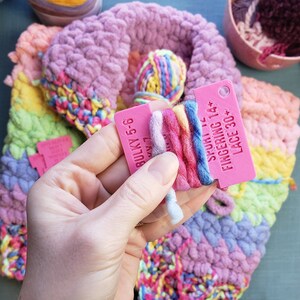 Op de afbeelding: Een close-up van kleurrijk garen en een lopend haakproject. Een roze label toont informatie over het gewicht van het garen. Het project heeft een verscheidenheid aan pastelkleuren, waaronder roze, geel en blauw. Een haaknaald is zichtbaar.