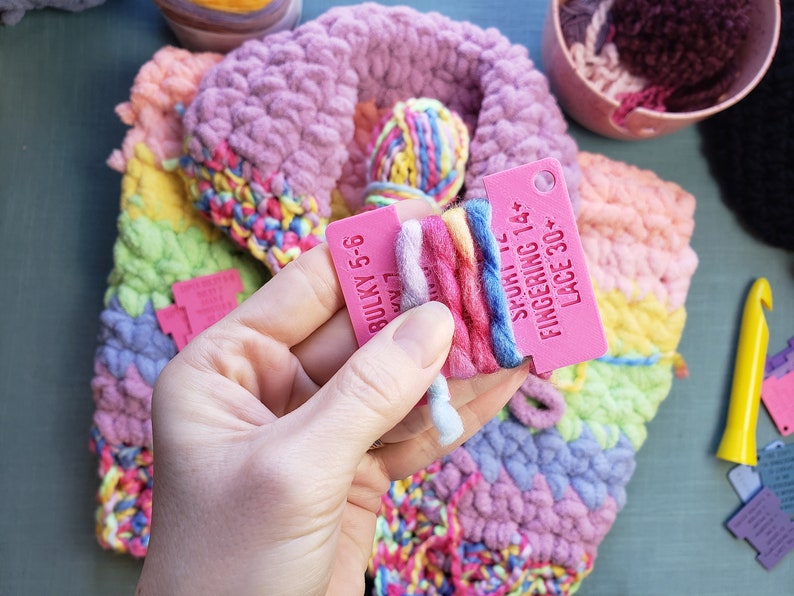 Op de afbeelding: Een close-up van kleurrijk garen en een lopend haakproject. Een roze label toont informatie over het gewicht van het garen. Het project heeft een verscheidenheid aan pastelkleuren, waaronder roze, geel en blauw. Een haaknaald is zichtbaar.