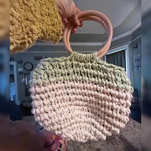 May include: A handmade crochet bag with a two-tone design. The top section is light green, transitioning to a soft pink at the bottom. It features two wooden handles. The bag is held up by a person.