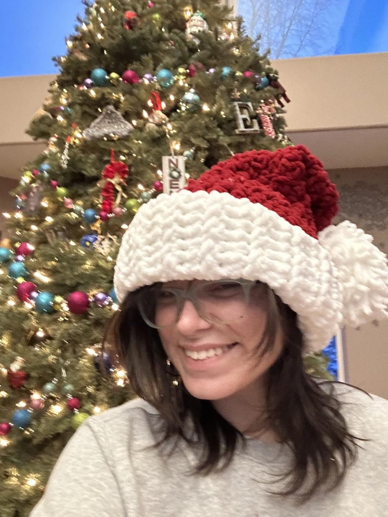 Op de afbeelding: Een vrouw draagt een witte en rode gebreide kerstmanmuts met een pluche witte pompon. Ze glimlacht en kijkt naar de camera. Op de achtergrond staat een kerstboom met kleurrijke kerstballen.
