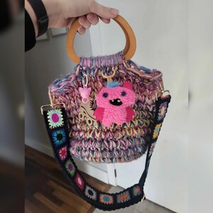May include: A handmade, multi-colored crochet bag with a wooden handle and a detachable strap. The bag features a pink monster charm and a roller skate charm. The strap is black with colorful floral crochet squares.