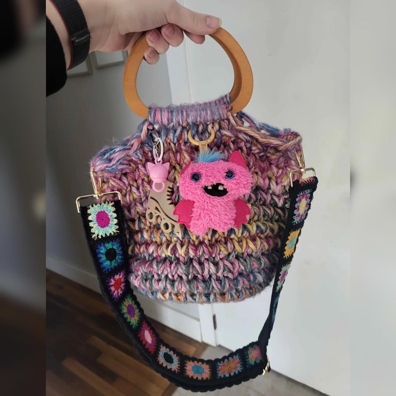 May include: A handmade, multi-colored crochet bag with a wooden handle and a detachable strap. The bag features a pink monster charm and a roller skate charm. The strap is black with colorful floral crochet squares.
