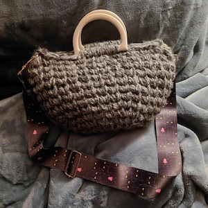 May include: A gray crocheted handbag with a white handle and a patterned shoulder strap. The strap features a repeating design of black cat silhouettes with pink hearts and yellow dots. The bag is sitting on a blue surface.