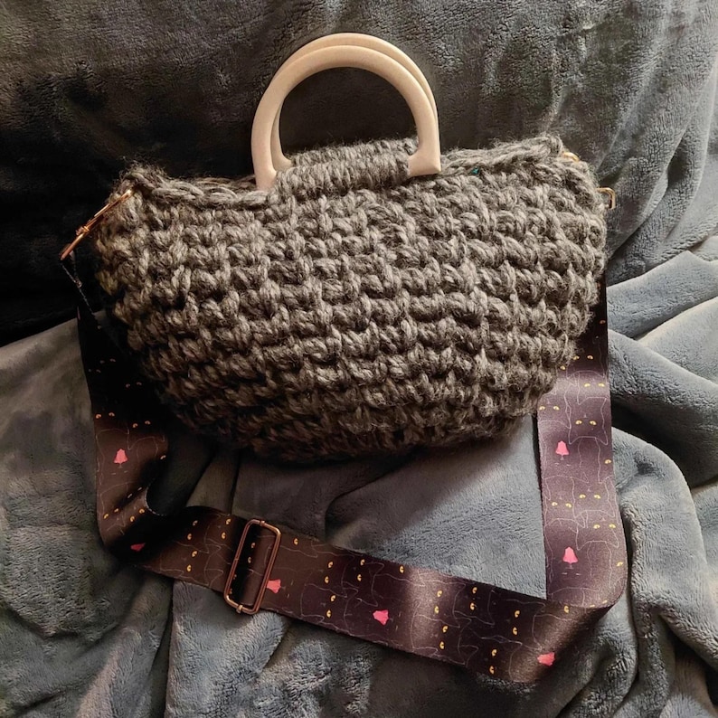 May include: A gray crocheted handbag with a white handle and a patterned shoulder strap. The strap features a repeating design of black cat silhouettes with pink hearts and yellow dots. The bag is sitting on a blue surface.