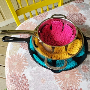 May include: A set of three crocheted pot holders in pink, yellow, and teal, stacked on top of a black cast iron skillet.