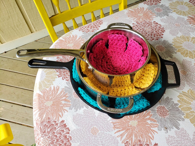 May include: A set of three crocheted pot holders in pink, yellow, and teal, stacked on top of a black cast iron skillet.