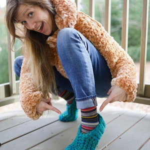 May include: A woman wearing a brown fuzzy jacket and blue jeans is squatting down. She is wearing colorful striped socks and teal crocheted slippers.