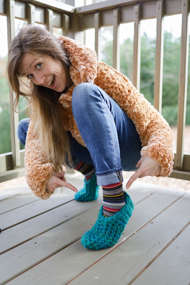May include: A woman wearing a brown fuzzy jacket and blue jeans is squatting down. She is wearing colorful striped socks and teal crocheted slippers.
