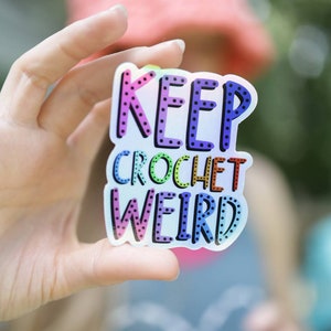 May include: A colorful sticker with the text "Keep Crochet Weird" in a rainbow gradient with black polka dots.