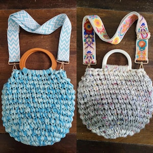 May include: Two round woven handbags with decorative straps. One is light blue with a wooden handle and a light blue and white patterned strap. The other is gray with a white handle and a floral strap.