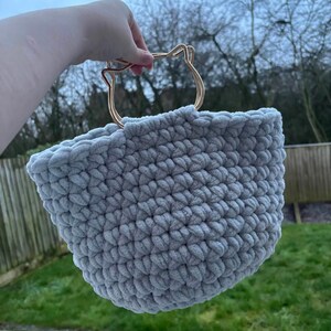 May include: A light gray, hand-crocheted basket with a gold-colored cat-shaped handle. The basket is made of a soft, plush yarn and has a rounded shape. The handle is a decorative element, adding a unique touch to the design. The basket is held up against a blurred outdoor background.