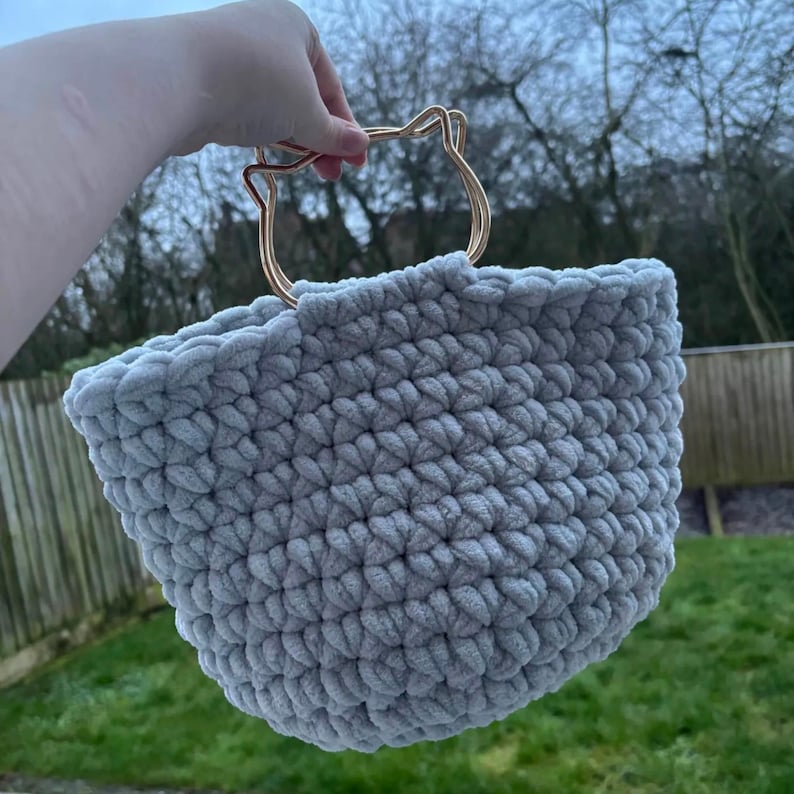 May include: A light gray, hand-crocheted basket with a gold-colored cat-shaped handle. The basket is made of a soft, plush yarn and has a rounded shape. The handle is a decorative element, adding a unique touch to the design. The basket is held up against a blurred outdoor background.