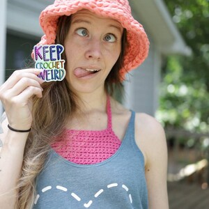 May include: A woman wearing a pink crocheted hat and a blue tank top with the text "CRAFTY" on it. She is holding a sticker that says "KEEP CROCHET WEIRD" in rainbow colors.