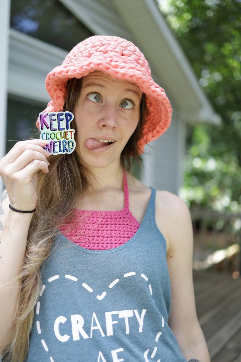 May include: A woman wearing a pink crocheted hat and a blue tank top with the text "CRAFTY" on it. She is holding a sticker that says "KEEP CROCHET WEIRD" in rainbow colors.