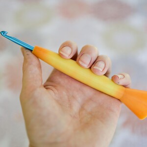 May include: A crochet hook with a blue metal hook and a yellow and orange handle. The hook is held in a hand against a blurred background. The handle is a gradient of yellow to orange.