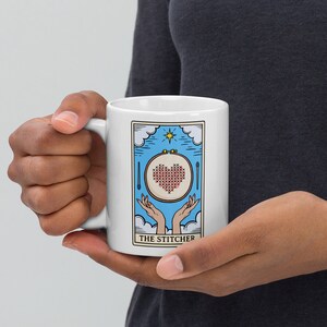 May include: White ceramic mug with a blue and white illustration of a hand-stitched heart in a hoop. The text "THE STITCHER" is printed below the illustration.