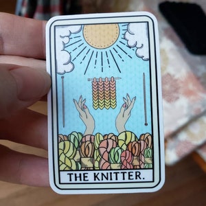 May include: A sticker featuring a tarot card design with the text "THE KNITTER." The card depicts a sun, clouds, knitting needles, hands, and yarn. The color palette includes light blue, yellow, and various shades of yarn.
