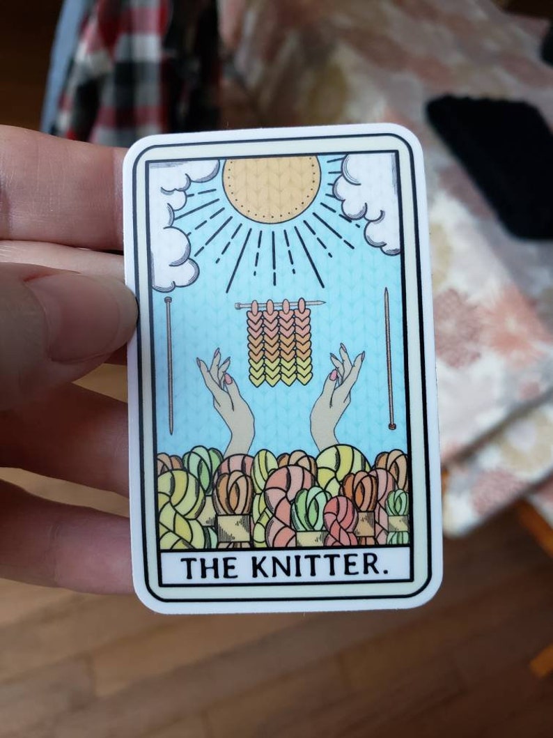 May include: A sticker featuring a tarot card design with the text "THE KNITTER." The card depicts a sun, clouds, knitting needles, hands, and yarn. The color palette includes light blue, yellow, and various shades of yarn.
