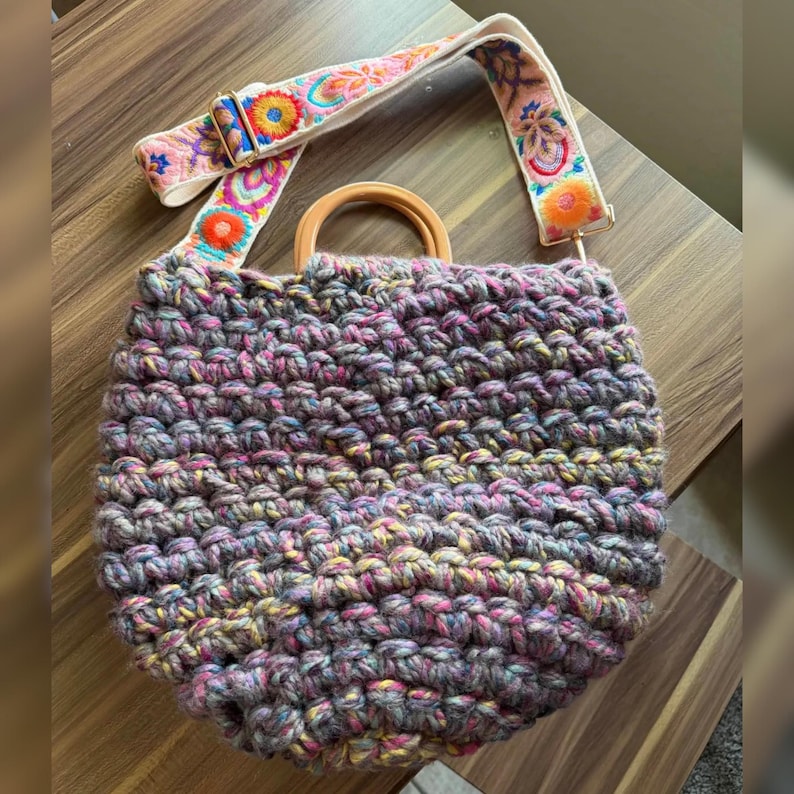May include: A handmade crochet bag with a colorful, textured design. The bag features a wooden handle and a decorative shoulder strap with floral embroidery in pink, orange, and blue. The bag's body is a mix of gray, pink, and yellow yarn.
