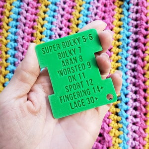 Op de afbeelding: Een felgroene garenmeter met tekst die garendiktes aangeeft van Super Bulky 5-6 tot Lace 30+. De meter wordt tegen een kleurrijke gehaakte achtergrond gehouden.