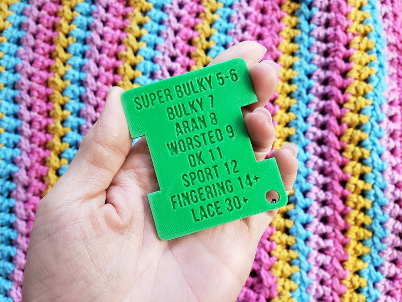 Op de afbeelding: Een felgroene garenmeter met tekst die garendiktes aangeeft van Super Bulky 5-6 tot Lace 30+. De meter wordt tegen een kleurrijke gehaakte achtergrond gehouden.