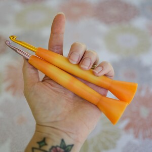 May include: Two crochet hooks with orange handles and metallic hook ends are held in a hand. One hook is gold, the other is silver. The handles have a gradient effect, transitioning from orange to yellow.