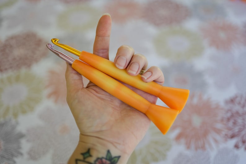 May include: Two crochet hooks with orange handles and metallic hook ends are held in a hand. One hook is gold, the other is silver. The handles have a gradient effect, transitioning from orange to yellow.