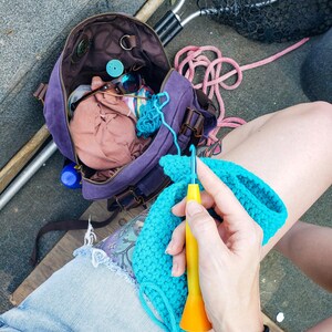 May include: A person is crocheting with a yellow and orange crochet hook and turquoise yarn. A purple bag with brown leather straps and a net are in the background. The bag contains sunglasses and other items.