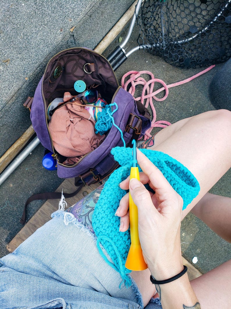 May include: A person is crocheting with a yellow and orange crochet hook and turquoise yarn. A purple bag with brown leather straps and a net are in the background. The bag contains sunglasses and other items.