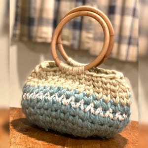 May include: A handmade crochet bag with a light blue and beige color scheme. The bag features a wooden handle and a white decorative stitch. The bag is sitting on a wooden surface.