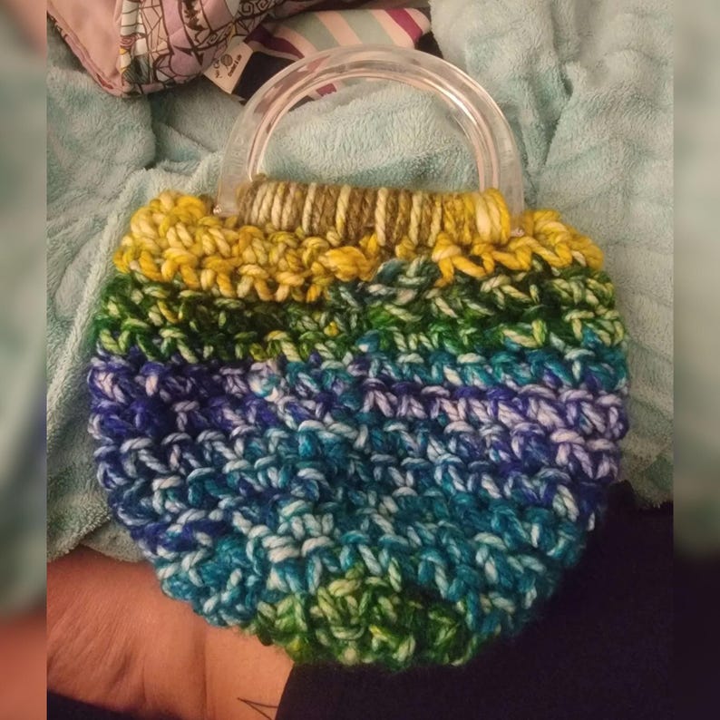 May include: A handmade crochet handbag with a clear acrylic handle. The bag features a vibrant color gradient, transitioning from yellow to green, blue, and teal. The yarn has a chunky texture, giving the bag a unique, textured appearance.