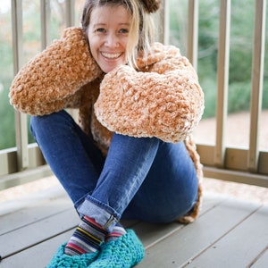 May include: A woman wearing a brown fuzzy sweater and blue jeans sits on a wooden deck. She is wearing teal blue crocheted slippers with colorful striped socks.