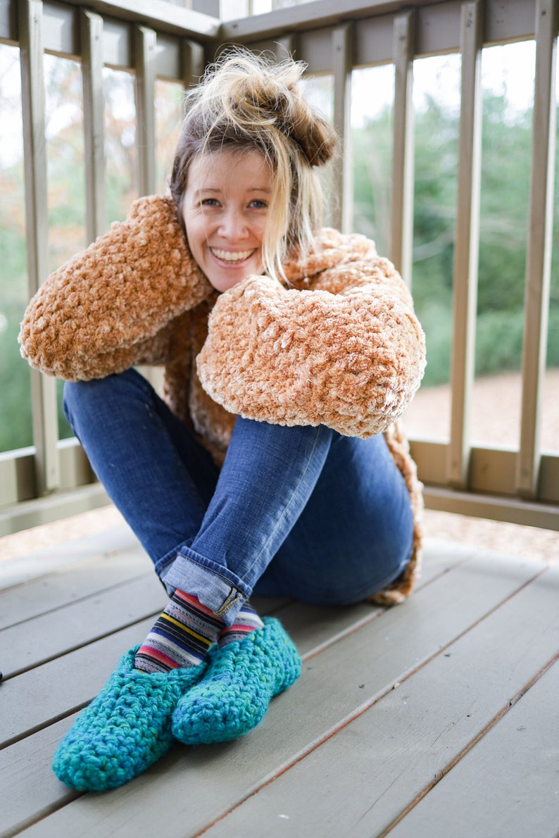 May include: A woman wearing a brown fuzzy sweater and blue jeans sits on a wooden deck. She is wearing teal blue crocheted slippers with colorful striped socks.