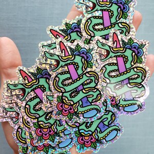 May include: A collection of holographic stickers featuring a green snake pierced by a purple dagger. The snake has an open mouth, red tongue, and is decorated with yellow and black details. Floral and leaf accents surround the snake.