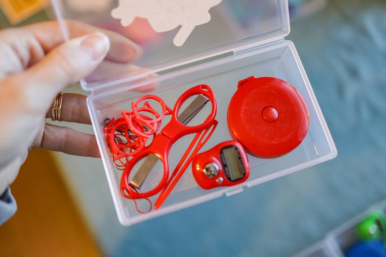 Pode incluir: Um recipiente de pl&aacute;stico transparente com uma fita m&eacute;trica vermelha, uma tesoura vermelha, el&aacute;sticos de cabelo vermelhos e um contador digital vermelho.