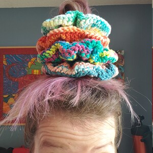 May include: A stack of colorful crocheted hair scrunchies in shades of blue, green, yellow, orange, and pink. The scrunchies are stacked on top of each other and are being worn by a person with pink hair.
