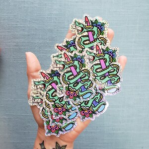 May include: A handful of holographic stickers featuring a green snake wrapped around a pink sword. The snake has a pink horn and is decorated with flowers and leaves. The stickers have a glittery, iridescent finish.