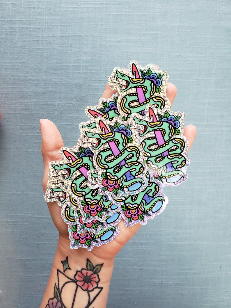 May include: A handful of holographic stickers featuring a green snake wrapped around a pink sword. The snake has a pink horn and is decorated with flowers and leaves. The stickers have a glittery, iridescent finish.