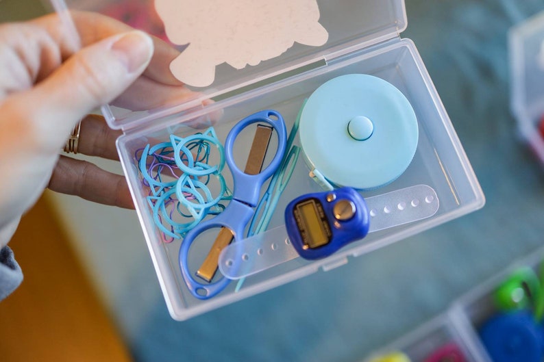 Pode incluir: Um recipiente de pl&aacute;stico transparente cheio de v&aacute;rios materiais de artesanato, incluindo clipes de papel azuis em forma de gato, um par de tesouras azuis, uma fita m&eacute;trica azul, um contador digital azul e um espeto de madeira.