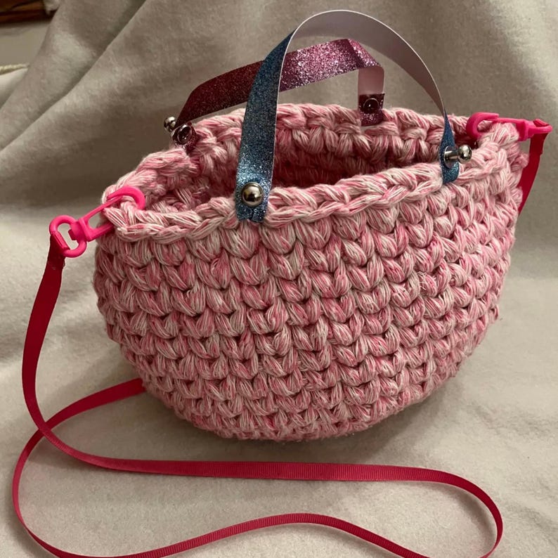 May include: A handmade pink crochet bag with a matching pink strap. The bag has a rounded shape and features a textured stitch pattern. The handles are made of white paper with glittery blue and red accents. The bag is secured with pink plastic clasps.