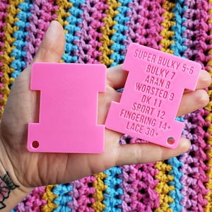 Op de afbeelding: Twee felroze garenmeetgereedschappen worden tegen een kleurrijke gehaakte achtergrond gehouden. E&eacute;n gereedschap heeft een reeks garen gewicht namen en nummers erop gedrukt, waaronder "SUPER BULKY 5-6" en "LACE 30+".