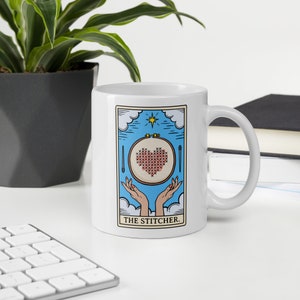 May include: White ceramic mug with a blue and white illustration of a hand holding a hoop with a red heart cross-stitched inside. The text "THE STITCHER." is printed below the illustration.