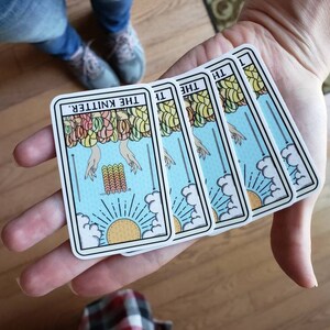 May include: Four tarot-style cards with a light blue background, featuring the text "THE KNITTER." The cards depict hands, yarn, and a sun. The cards are held in a person's hand.