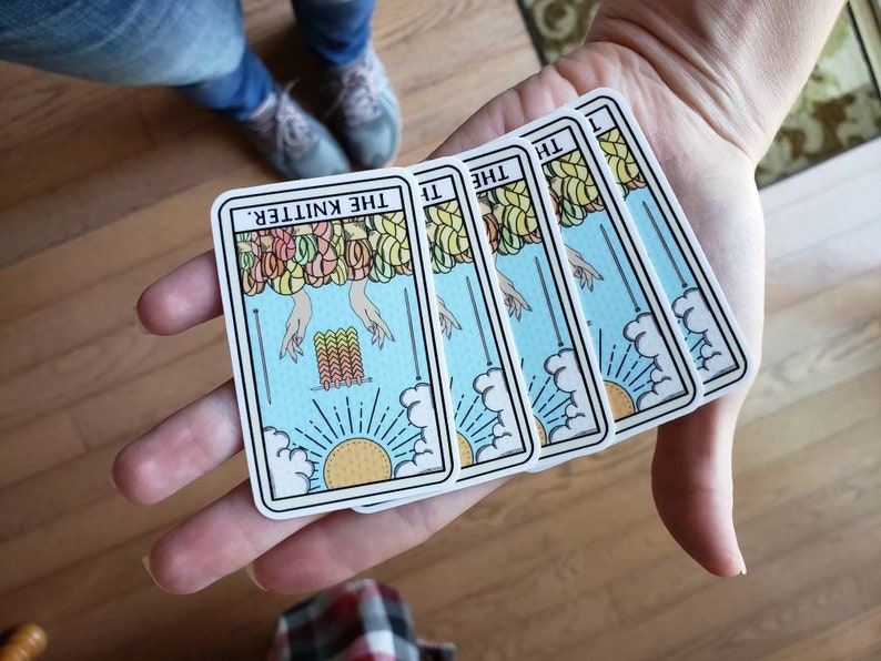 May include: Four tarot-style cards with a light blue background, featuring the text "THE KNITTER." The cards depict hands, yarn, and a sun. The cards are held in a person's hand.