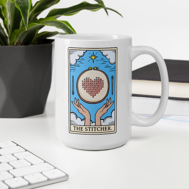 May include: White ceramic mug with a blue and white illustration of a hand holding a hoop with a red heart cross-stitched inside. The text "THE STITCHER." is printed below the illustration.