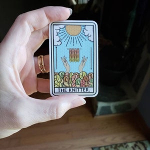 May include: A rectangular sticker featuring a tarot card design with a blue background, a sun, and hands knitting. The card reads "THE KNITTER" in black text. The sticker is held by a hand.