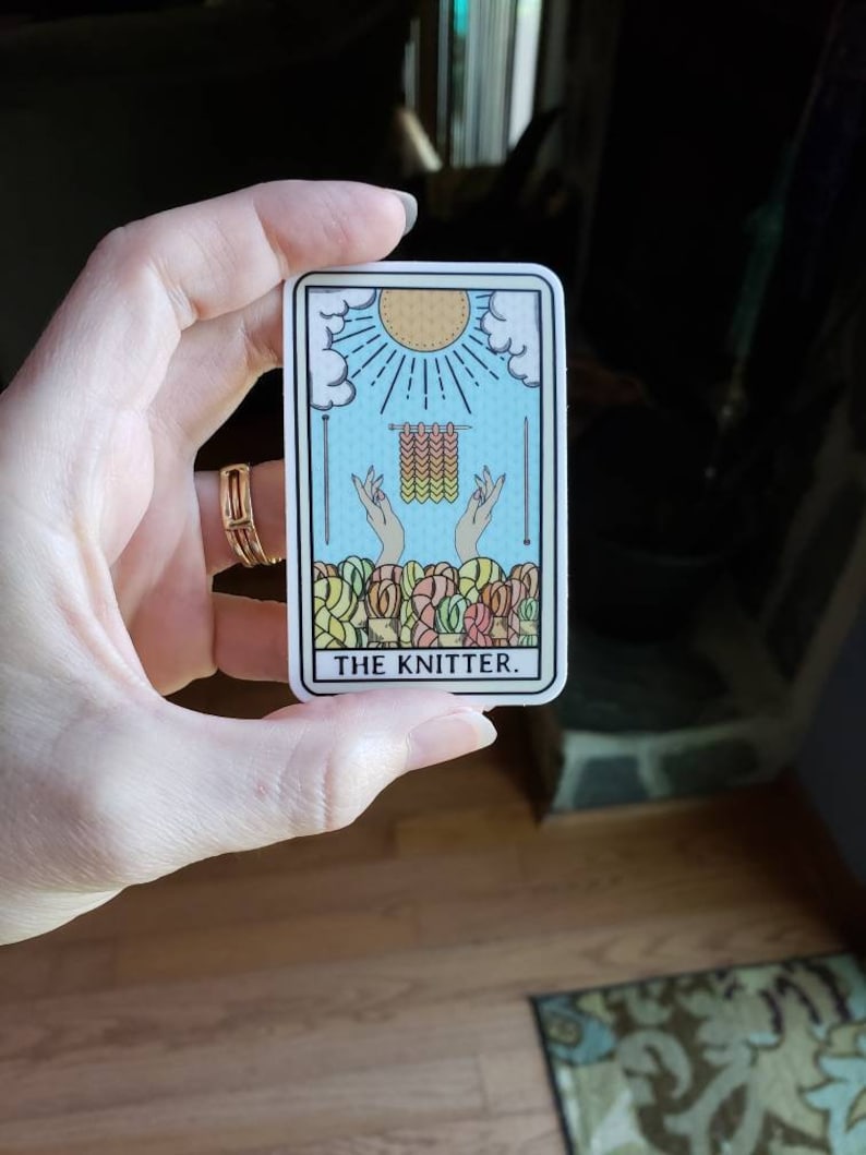 May include: A rectangular sticker featuring a tarot card design with a blue background, a sun, and hands knitting. The card reads "THE KNITTER" in black text. The sticker is held by a hand.