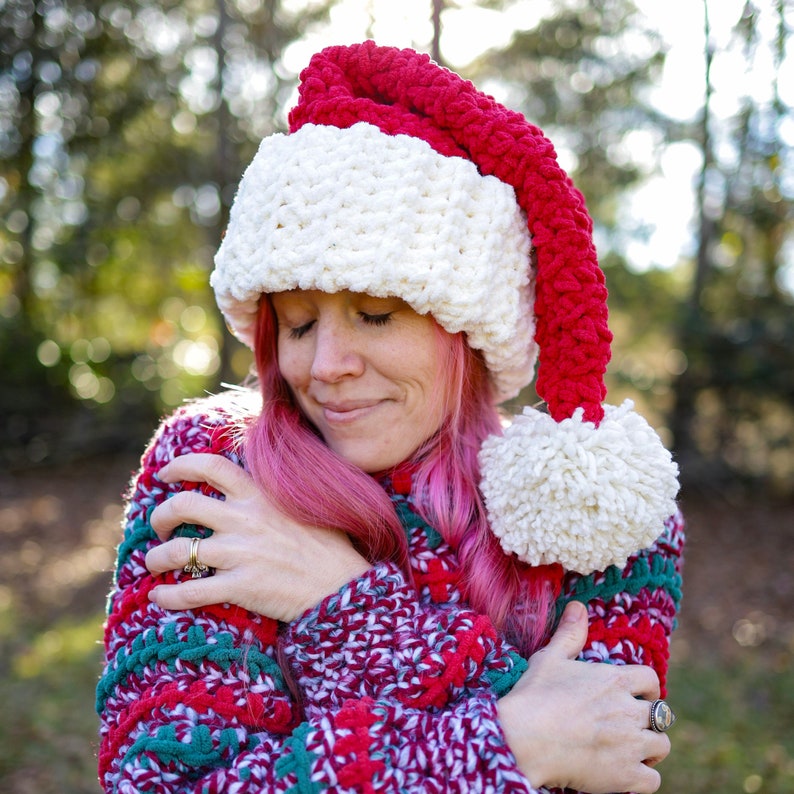 Op de afbeelding: Een vrouw draagt een witte en rode gebreide kerstmanmuts met een witte pompon. Ze draagt een rood, groen en wit gebreide trui en heeft roze haar.