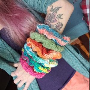 May include: A stack of colorful crocheted bracelets in various shades of pink, green, orange, blue, and white. The bracelets are made with a textured stitch and are stacked on top of each other.